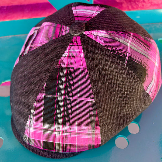 A structured flat cap with a dark brown herringbone fabric crown and bright pink, black, and white plaid panels, resting on a turquoise surface.