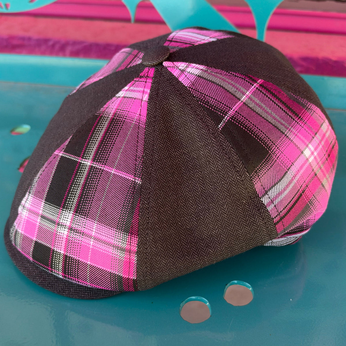 A six-panel flat cap with a dark brown twill base and a vibrant pink, black, and white plaid pattern on the front panels, resting on a turquoise metal surface.