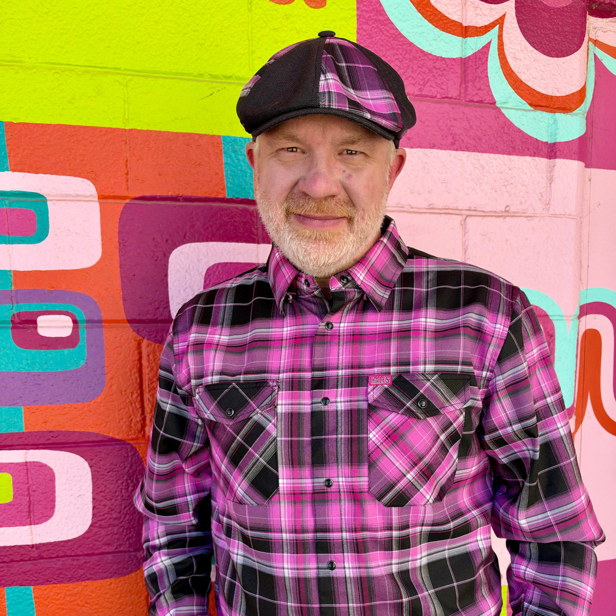 A man wearing a pink and black plaid button-down shirt and a matching flat cap, standing in front of a brightly colored geometric mural.