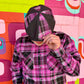 A person wearing a pink, black, and white plaid button-up shirt and a matching plaid cap, posing in front of a vibrant, colorful geometric brick wall.