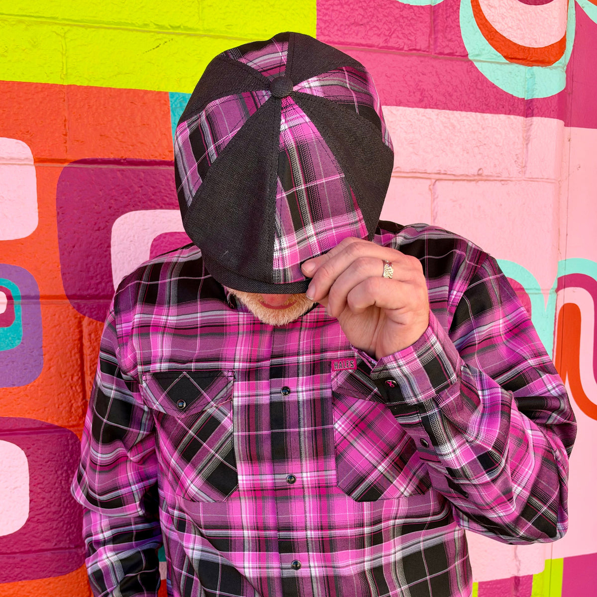 A person wearing a pink, black, and white plaid button-up shirt and a matching plaid cap, posing in front of a vibrant, colorful geometric brick wall.