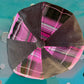 A top-down view of a structured flat cap with alternating panels of dark brown herringbone fabric and bright pink plaid fabric, resting on a turquoise metal surface.