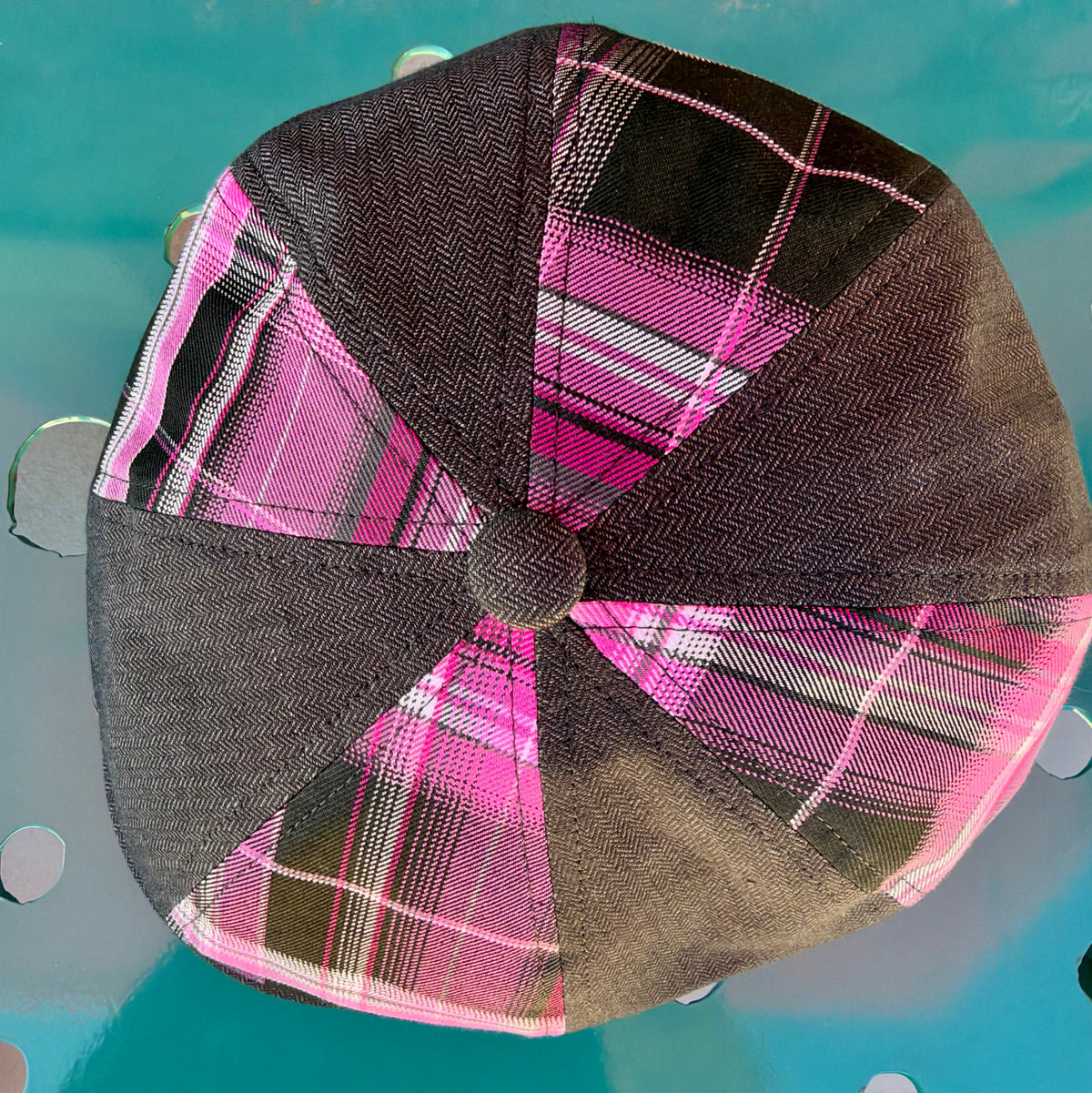 A top-down view of a structured flat cap with alternating panels of dark brown herringbone fabric and bright pink plaid fabric, resting on a turquoise metal surface.