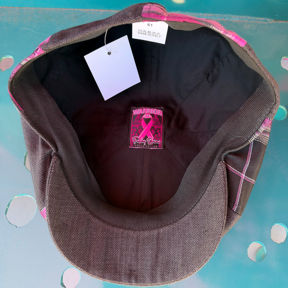 A dark brown textured fabric newsboy cap with pink and white plaid trim, shown from the inside. The cap features a black interior, a pink ribbon patch on the crown, and a white size tag.