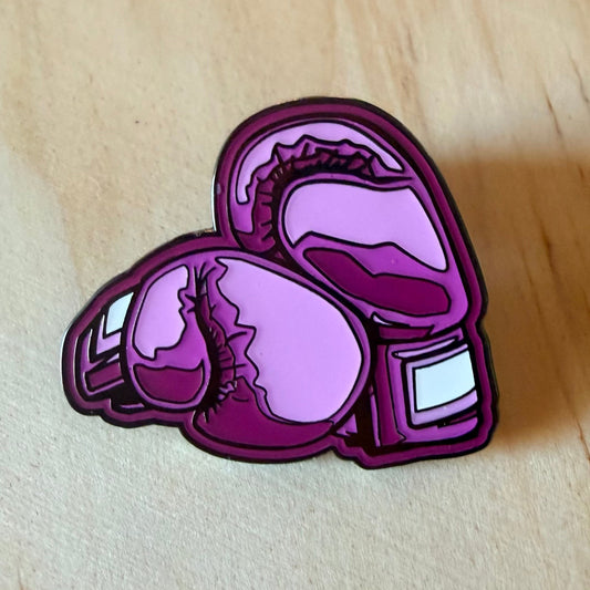 A purple and pink boxing gloves enamel pin with black outlines, displayed on a light wood background.