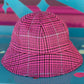 A pink and black houndstooth bucket hat with a subtle grid pattern, resting on a colorful teal and pink surface.