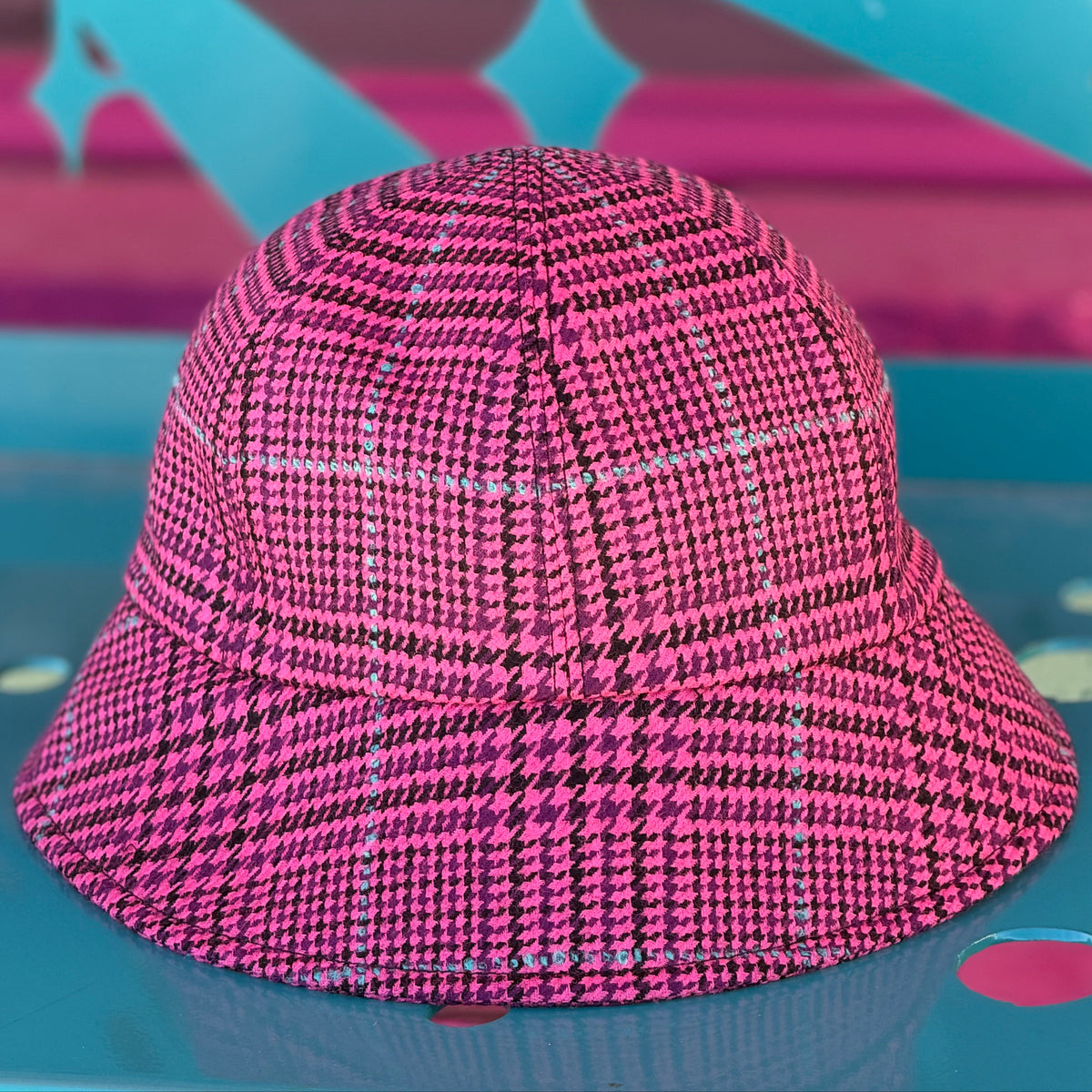 A pink and black houndstooth bucket hat with a subtle grid pattern, resting on a colorful teal and pink surface.