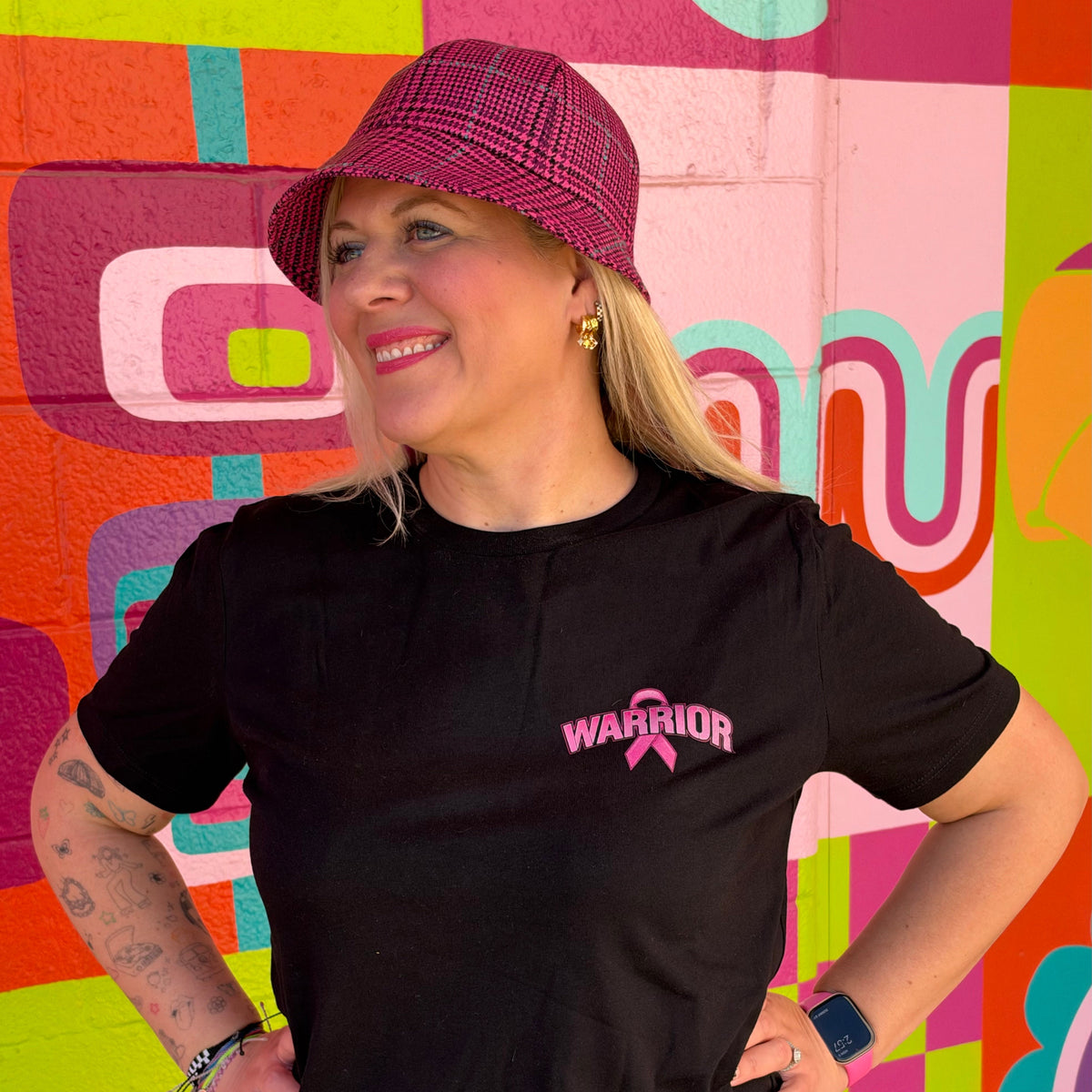 A woman wearing a black short-sleeve t-shirt with a pink 'WARRIOR' and pink ribbon graphic on the left chest, paired with a pink plaid bucket hat and blue jeans, standing in front of a colorful geometric mural.