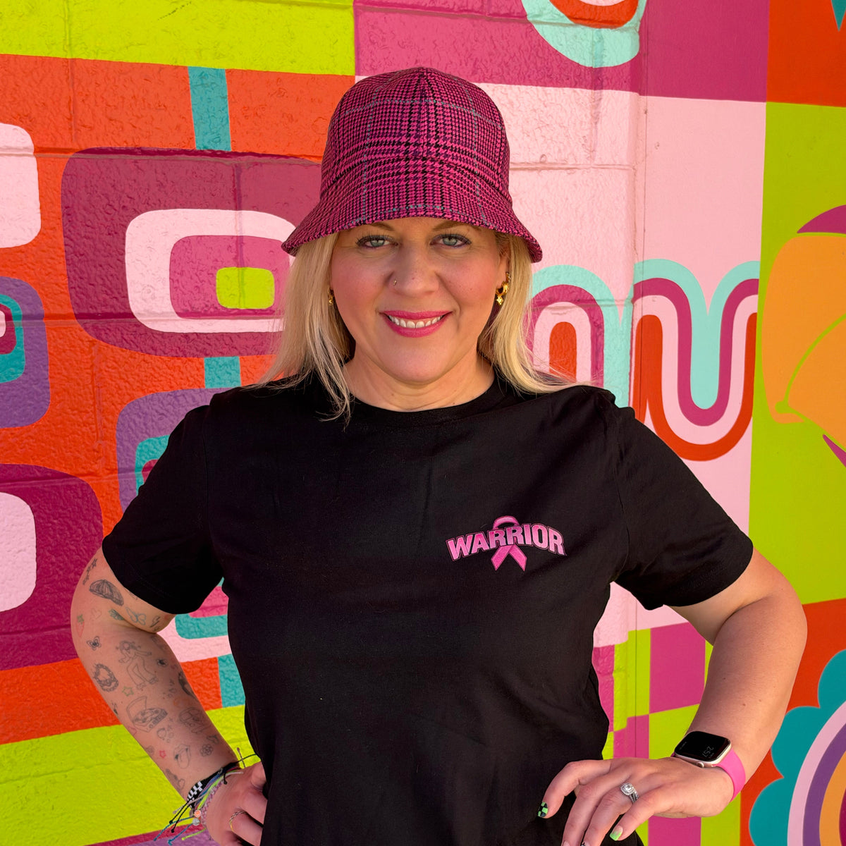 A woman wearing a black short-sleeve t-shirt with a pink 'WARRIOR' ribbon graphic on the left chest, paired with a dark pink plaid bucket hat and blue denim shorts, standing in front of a colorful graffiti wall.