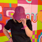 A person wearing a pink and purple plaid bucket hat and a black t-shirt stands in front of a brightly colored geometric mural wall.