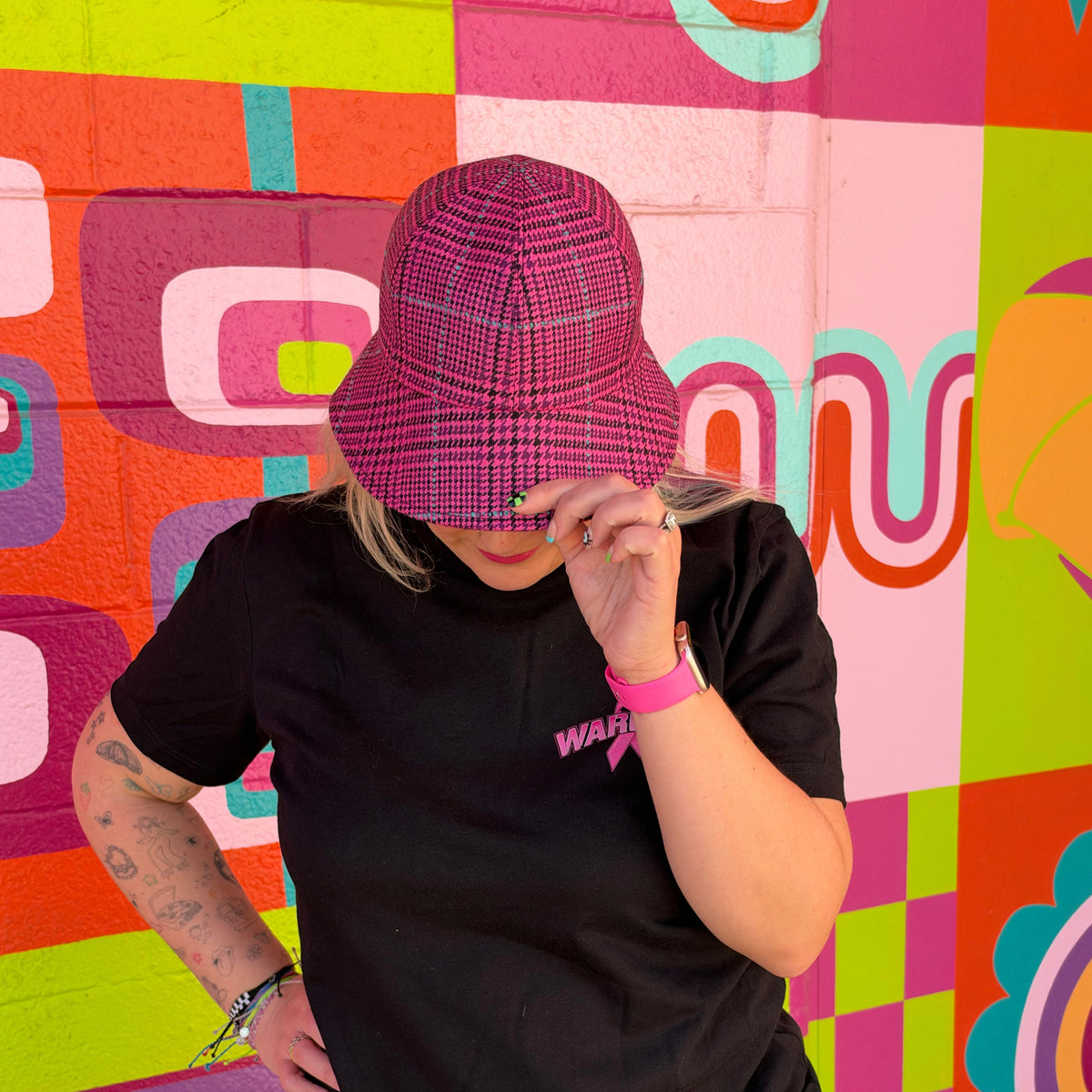 A person wearing a pink and purple plaid bucket hat and a black t-shirt stands in front of a brightly colored geometric mural wall.