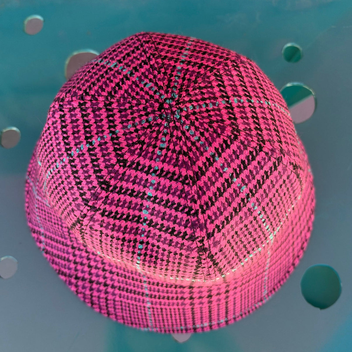A pink and black patterned hexagonal kippah or skullcap photographed from above against a teal-blue background with circular cutouts.