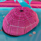 A bright pink and black patterned dome-shaped hat or cover with a small top hole, photographed on a glossy turquoise surface with colorful abstract background elements.