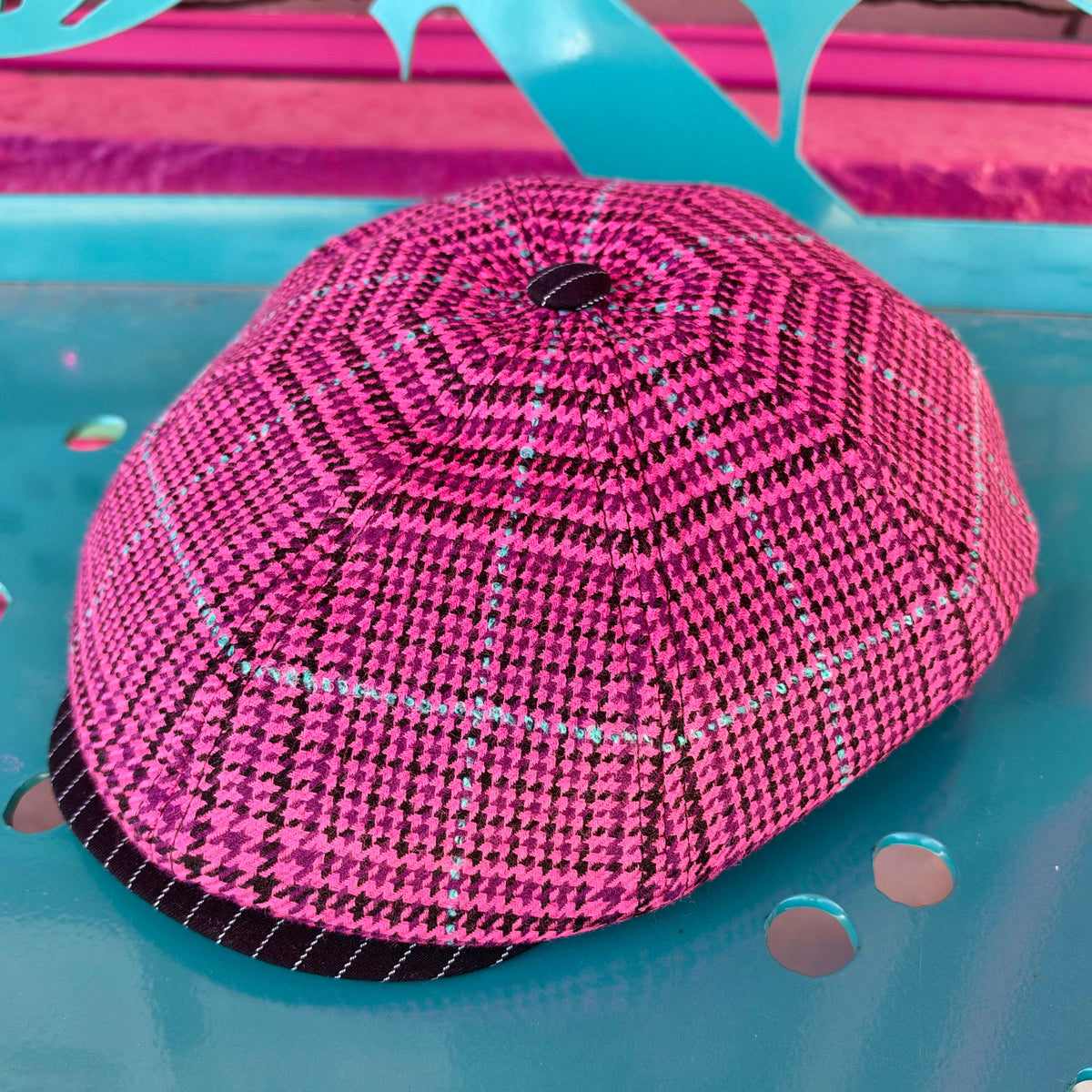 A bright pink and black patterned dome-shaped hat or cover with a small top hole, photographed on a glossy turquoise surface with colorful abstract background elements.