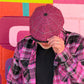 A person wearing a pink and black plaid flat cap and a matching pink and black plaid button-up shirt over a black t-shirt, standing in front of a colorful geometric wall.