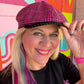 A smiling woman wearing a pink and black houndstooth flat cap, posing in front of a colorful graffiti wall. She is wearing a black t-shirt with pink lettering.
