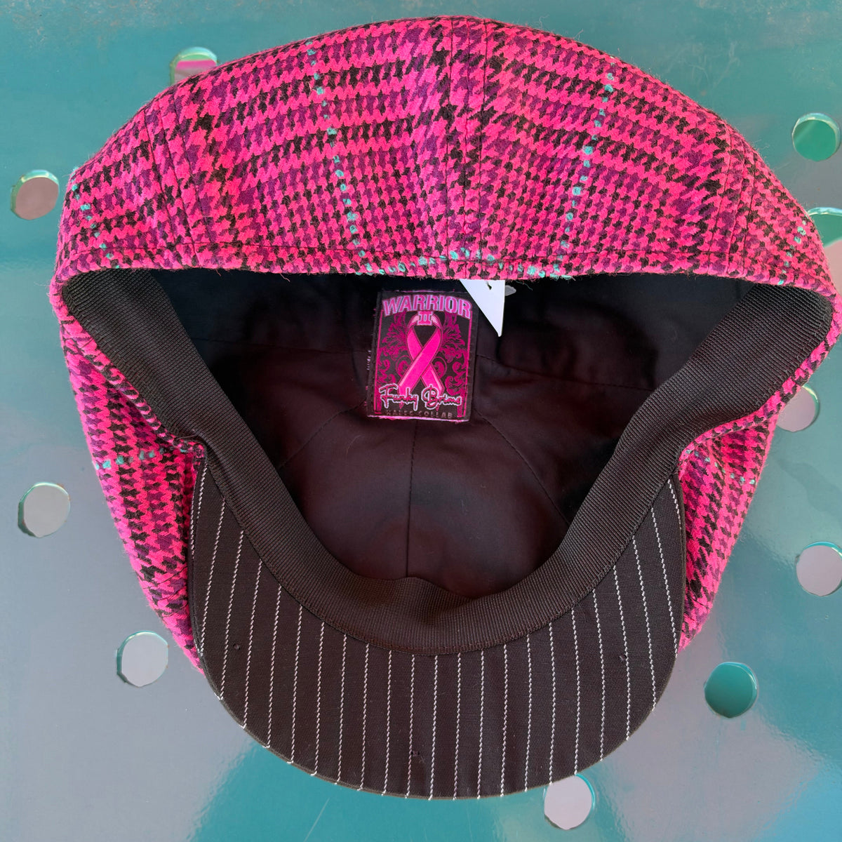 A pink and black patterned flat cap with a brown pinstriped brim, photographed from the inside. The interior lining is brown, and a pink ribbon patch is visible on the inner crown.